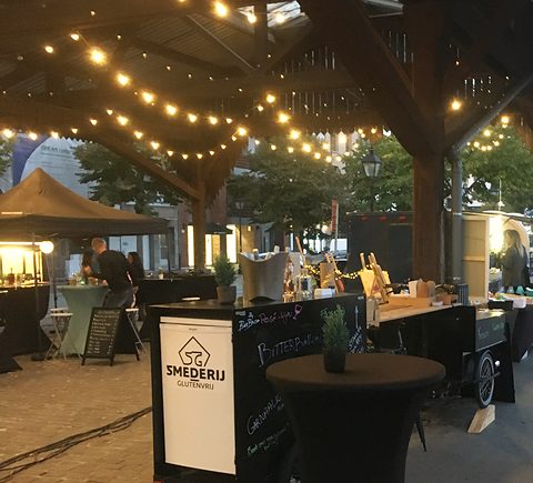 Het kraam van Smederij Glutenvrij op een markt bij valavond, onder lichtslingers
