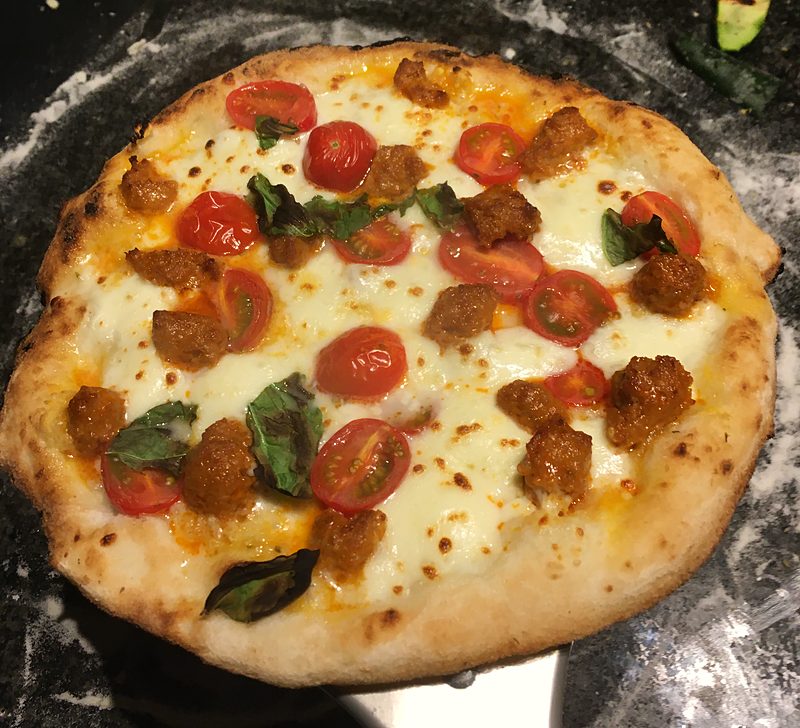 Pizza met gesmolten mozzarella, kerstomaatjes, stukjes chorizogehakt en basilicum, op een pizzaschep met bloem